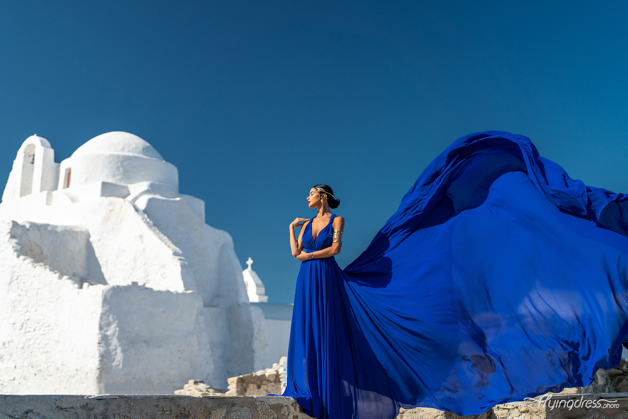 A woman in a flowing blue dress poses elegantly against the backdrop of the Paraportiani Church in Mykonos, with her dress billowing dramatically in the wind and a clear blue sky overhead.